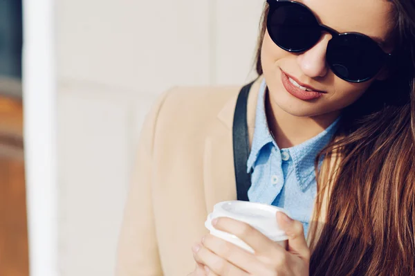 Menina bonita e jovem em um casaco e cachecol e óculos de sol sentados no banco. Mulher a beber café. Verão . — Fotografia de Stock