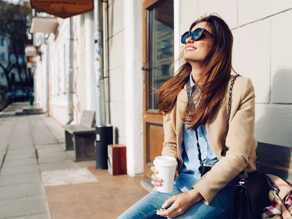 Hermosa y joven chica en un abrigo y bufanda y gafas de sol sentado en el banco. Mujer bebiendo café y riendo. Verano . — Foto de Stock