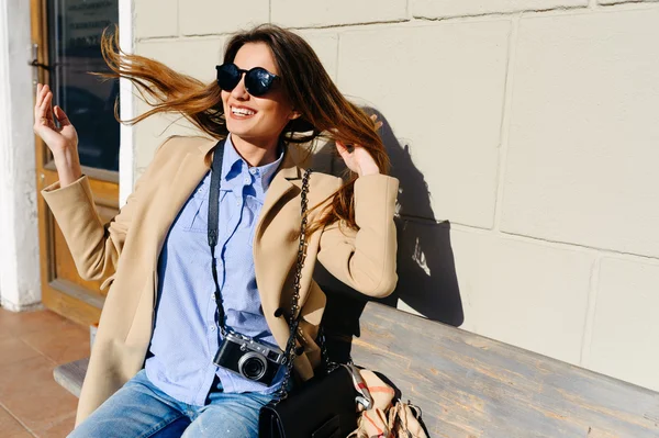 Hermosa y joven chica en un abrigo y bufanda y gafas de sol sentado en el banco y se ríe. Verano. El sol — Foto de Stock