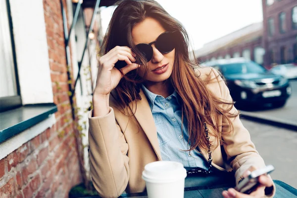 Bella e giovane ragazza che riposa in un caffè estivo. Donna che beve caffè e scrive un messaggio sul suo telefono — Foto Stock