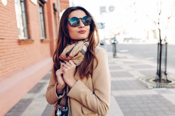 Hermosa y joven chica en un abrigo y bufanda y gafas de sol caminando por la ciudad en el verano . — Foto de Stock