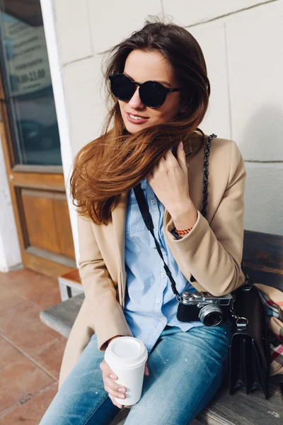 Hermosa y joven chica en un abrigo y bufanda y gafas de sol sentado en el banco. Mujer bebiendo café. Verano . — Foto de Stock