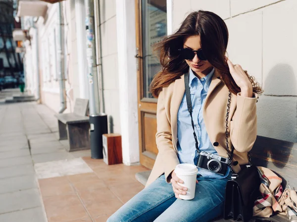 Menina bonita e jovem em um casaco e cachecol e óculos de sol sentados no banco. Mulher a beber café. Verão . — Fotografia de Stock