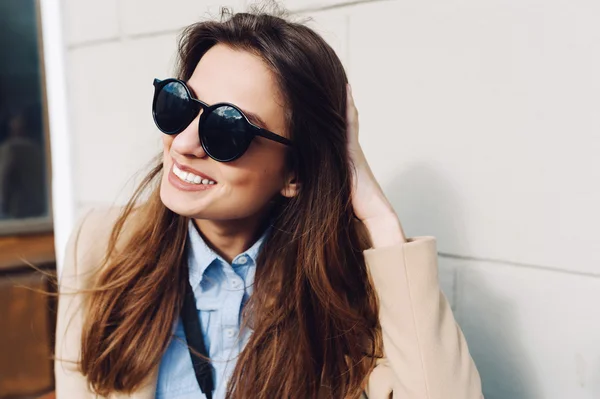 Bella e giovane ragazza in un cappotto e sciarpa e occhiali da sole seduti sulla panchina e ride. Estate. Il sole — Foto Stock