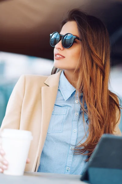 Красива і молода дівчина в сонцезахисних окулярах п'є каву на вулиці і читає новини в планшеті — стокове фото