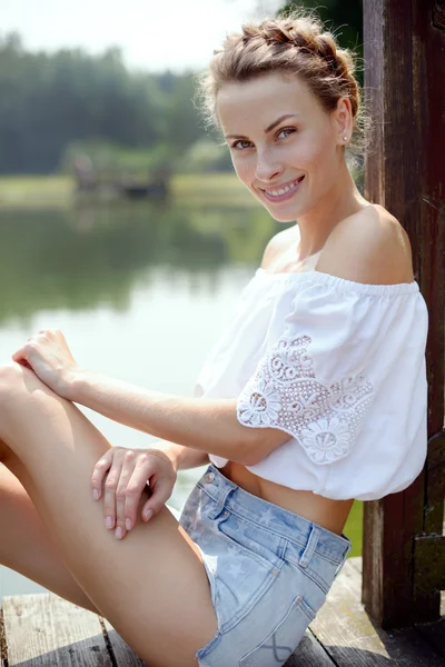 Young beautiful girl sitting on the beach near the lake in the summer in shorts and smiling — Stock Photo, Image