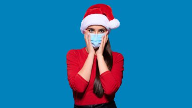 Heyecanlı kadın Noel Baba şapkası takıyor ve koruyucu tıbbi maske takıyor. Dokunaklı yüz, mavi izole arka plan..