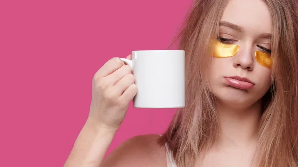 Mulher cansada e bêbada com ressaca e dor de cabeça colocar manchas nos olhos segurando xícara branca de café, isolar fundo rosa — Fotografia de Stock