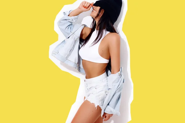 Slender tanned young woman with strong body in a denim jacket and shorts with a hat on a white wall