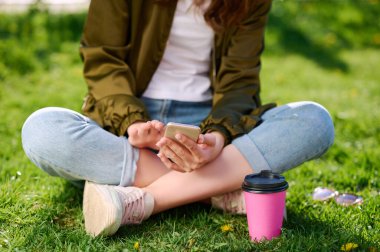 Şehir parkında çimlerde oturan ve açık havada akıllı telefon kullanan genç bir kız öğrenci.