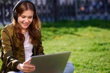 Kız öğrenci dışarıda çalışırken dizüstü bilgisayar kullanıyor ve yaz şehrinde kulaklıkla müzik dinliyor.
