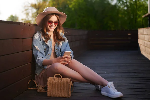 Hasır şapkalı gülümseyen kadın yaz kıyafetiyle dışarıda akıllı telefon kullanıyor ve ahşap terasta oturuyor. — Stok fotoğraf