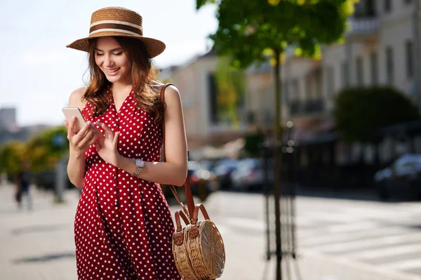 Güzel genç bir kadın akıllı telefon kullanıyor ve yaz şehrinde yürüyor. Hasır şapka ve benekli güzel kırmızı bir elbise giyiyor. — Stok fotoğraf
