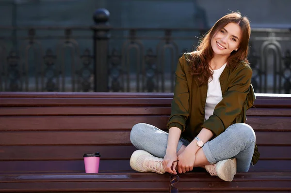 Mujer atractiva sonriente sentada en el banco en la ciudad de verano bebe taza de café caliente y disfruta de la luz del sol y el clima — Foto de Stock