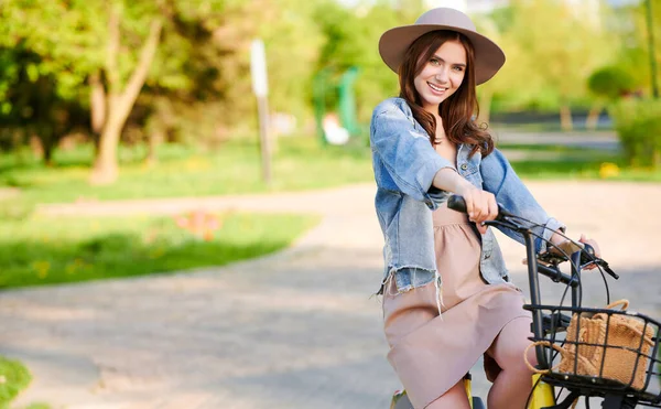 Woman in hat standing and smiling with bicycle outdoors in summer city — Stock Photo, Image