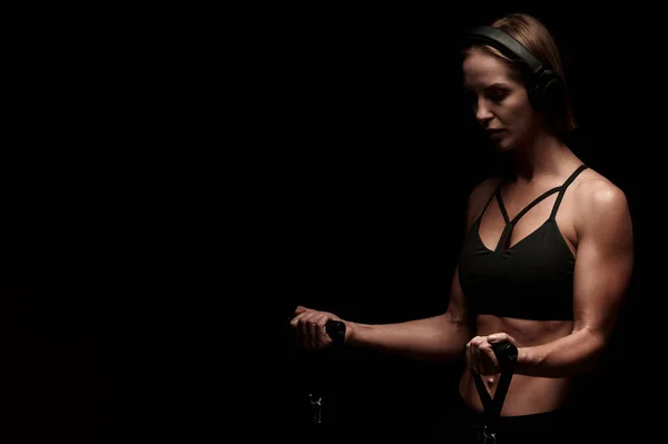 Sportswoman trénink s odporovými kapelami na černém pozadí a poslech hudby ve sluchátkách — Stock fotografie