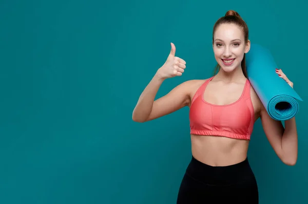 Joven mujer sonriente deportiva en ropa deportiva sosteniendo la esterilla de yoga en sus manos y mostrar el gesto de pulgar hacia arriba — Foto de Stock