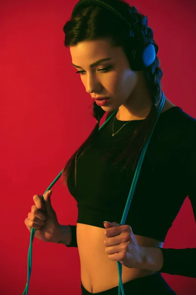 Mujer deportiva fuerte joven con cuerpo en forma escuchando música en auriculares y sosteniendo la cuerda de salto sobre fondo rojo —  Fotos de Stock