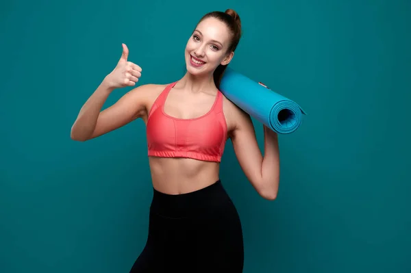 Joven mujer sonriente deportiva en ropa deportiva sosteniendo la esterilla de yoga en sus manos y mostrar el gesto de pulgar hacia arriba — Foto de Stock
