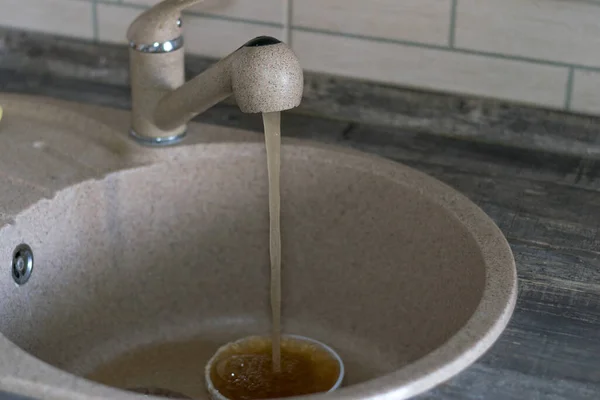 Agua oxidada sucia que fluye de la red. Grifo con agua mala en la casa en la cocina. El agua marrón anaranjada se recoge en una taza blanca. Ecología deficiente, medio ambiente contaminado, falta de agua limpia. Fotos De Stock