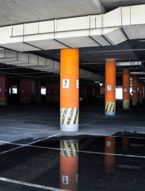 Binanın altına park etmek (otopark altı). Sayılı beton sütunlar. Asfalttaki su birikintileri park yerinin duvarlarını ve tavanını yansıtıyor..