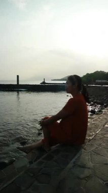 Turuncu gömlekli kadın, bulutlu bir günde beton çıkıntıda oturmuş kollarını açmış sakin sulara bakıyor..