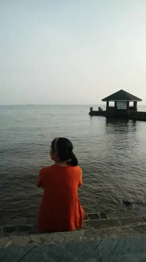 Turuncu gömlekli kadın, bulutlu bir günde beton çıkıntıda oturmuş kollarını açmış sakin sulara bakıyor..