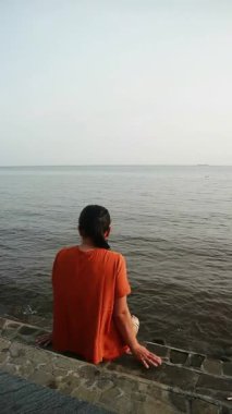 Turuncu gömlekli kadın, bulutlu bir günde beton çıkıntıda oturmuş kollarını açmış sakin sulara bakıyor..