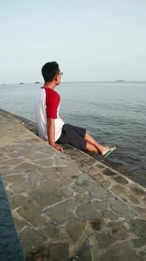 Genç adam sakin suların yanında, uzak bir manzarayla taş iskelede oturuyor.