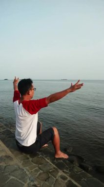 Genç adam sakin suların yanında, uzak bir manzarayla taş iskelede oturuyor.