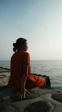 Turuncu gömlekli kadın, bulutlu bir günde beton çıkıntıda oturmuş kollarını açmış sakin sulara bakıyor..