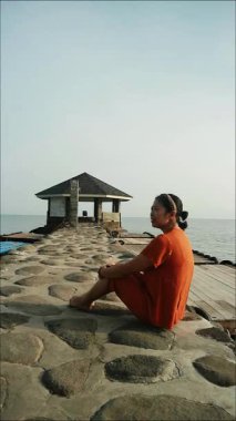 Turuncu gömlekli kadın bulutlu bir günde sakin su sahiline bakan Stoney Rıhtımı 'nda oturuyor, iyileşmek için seyahat ediyor.