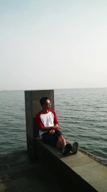 Sakin bir plajda okyanus esintisini kucaklayan Asyalı adam, zihinsel sağlık farkındalığı, tropikal turizm broşürleri ve yaşam tarzı bloglarında kullanmak için kişisel iyileşme ve solo seyahat anı yakalıyor.