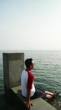 Sakin bir plajda okyanus esintisini kucaklayan Asyalı adam, zihinsel sağlık farkındalığı, tropikal turizm broşürleri ve yaşam tarzı bloglarında kullanmak için kişisel iyileşme ve solo seyahat anı yakalıyor.