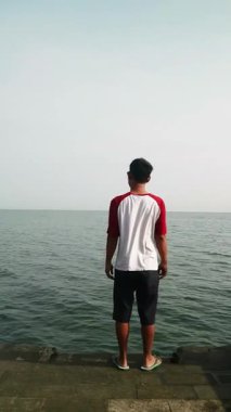 Sakin bir plajda okyanus esintisini kucaklayan Asyalı adam, zihinsel sağlık farkındalığı, tropikal turizm broşürleri ve yaşam tarzı bloglarında kullanmak için kişisel iyileşme ve solo seyahat anı yakalıyor.