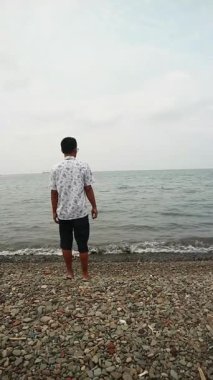 Sakin bir plajda okyanus esintisini kucaklayan Asyalı adam, zihinsel sağlık farkındalığı, tropikal turizm broşürleri ve yaşam tarzı bloglarında kullanmak için kişisel iyileşme ve solo seyahat anı yakalıyor.