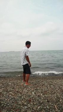 Sakin bir plajda okyanus esintisini kucaklayan Asyalı adam, zihinsel sağlık farkındalığı, tropikal turizm broşürleri ve yaşam tarzı bloglarında kullanmak için kişisel iyileşme ve solo seyahat anı yakalıyor.