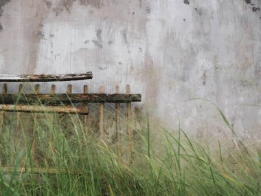 Yıpranmış ahşap bir çit, eskimiş beton bir arka planda kırsal bir duvar kağıdıyla uzun otların arasında duruyor.