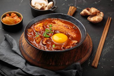 Yumurta sarısı, enoki, shiitake, tofu ve yeşil soğanlı, yemek çubukları ve yemek malzemeleriyle siyah bir kasede baharatlı kimçi jjigae çorbası. Kore mutfağı.