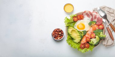 Healthy ketogenic diet concept showing a balanced meal on plate with salmon, avocado, fried egg, bacon, broccoli, tomato, and fresh salad, promoting low carb and high fat eating, copy space