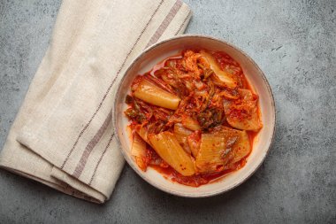Kimchi, mayalanmış bir Napa lahana kabağı, gri beton arka planda seramik bir kasede keten peçeteyle canlı rengini ve dokusunu yukarıdan aşağıya doğru görüntülüyor.
