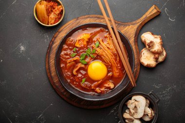 Yumurta sarısı, enoki, shiitake, tofu ve yeşil soğanlı, yemek çubukları ve yemek malzemeleriyle siyah bir kasede baharatlı kimçi jjigae çorbası. Kore mutfağı.