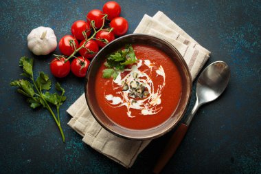 Taze domates çorbası krema, tohum ve otlarla dolu bir kasede servis edilir, yanında da mavi bir zemin üzerinde kaşık ve peçeteyle kiraz domatesleri, sarımsak ve maydanoz bulunur.