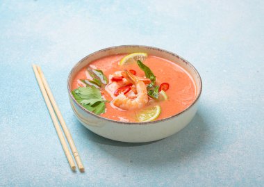 Taze karides, limon kamaları, şifalı otlar ve biberlerle süslenmiş ve yanında keten peçetede yemek çubukları bulunan Tayland 'ın popüler bir yemeği olan kremalı Tom yum çorbası bir kasede servis edilir.