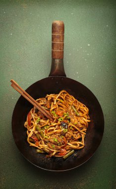 Tavada kızarmış tavuk ve sebzeli erişte, tepesinde tahta çubuklar, yeşil arka planda üst manzara.