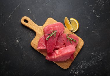 Taze kesilmiş ton balıklı biftekler, ahşap bir tahtanın üzerinde taze biberiye ve limon dilimleri ile tatlandırılmış pişirmeye hazırlanıyor.