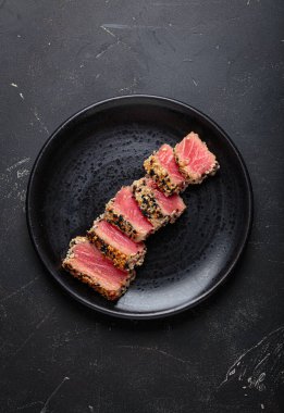 Kurutulmuş ton balığı dilimleri, siyah bir tabakta susam tohumlarıyla kaplı, yemek çubuklarıyla servis edilen, siyah taşlı bir arka planda.