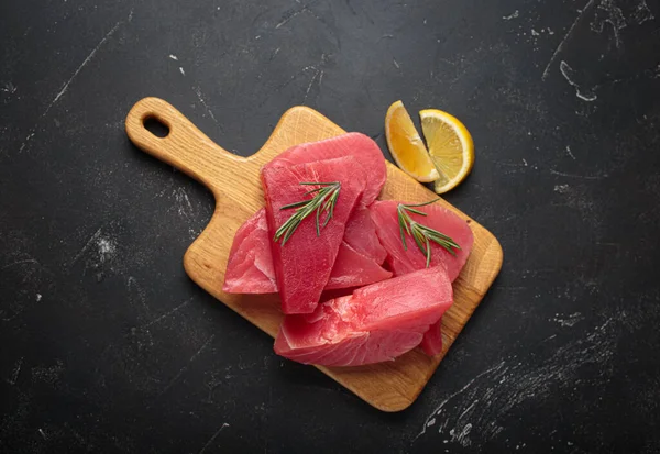 Taze kesilmiş ton balıklı biftekler, ahşap bir tahtanın üzerinde taze biberiye ve limon dilimleri ile tatlandırılmış pişirmeye hazırlanıyor.