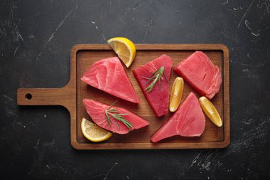 Taze kesilmiş ton balıklı biftekler, ahşap bir tahtanın üzerinde taze biberiye ve limon dilimleri ile tatlandırılmış pişirmeye hazırlanıyor.