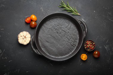 Taze sarımsak, domates, biberiye ve karışık biberiye ile çevrili koyu bir zemin üzerine demir tava dökmek